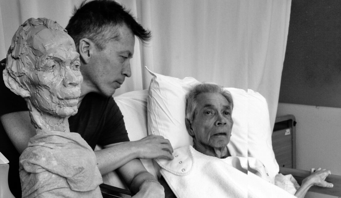 kleien beeld met op de achtergrond een oude man op een ziekenhuisbed