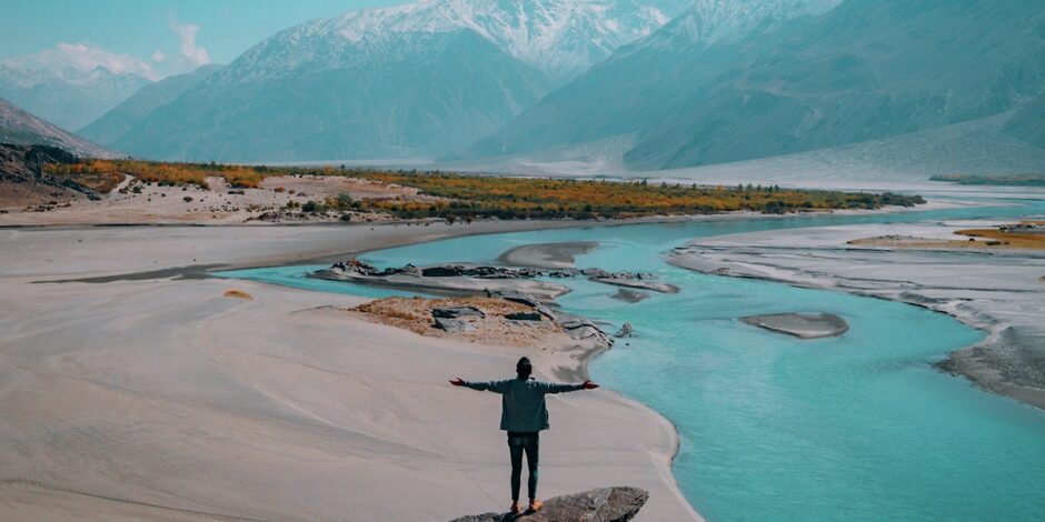 Man staat met zijn armen wijd naast een rivier met in de achtergrond bergen.