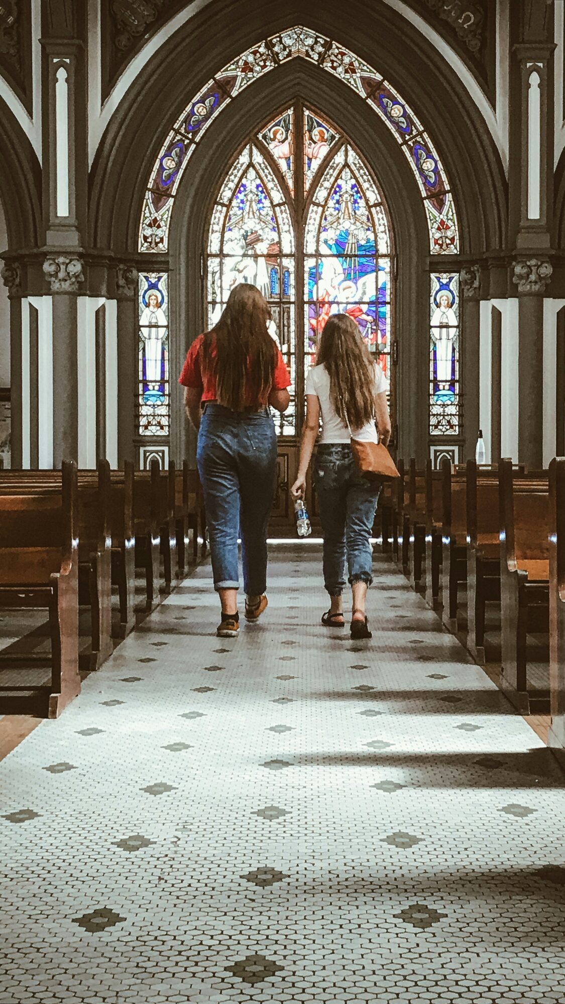 Twee lopende personen in een kerk met achter hen glas in lood ramen