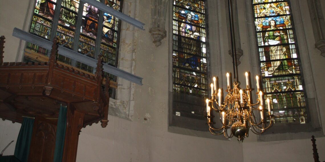 Binnenkant van een kerk. Links de preekstoel, rechts een kroonluchter met daarachter glas in lood.