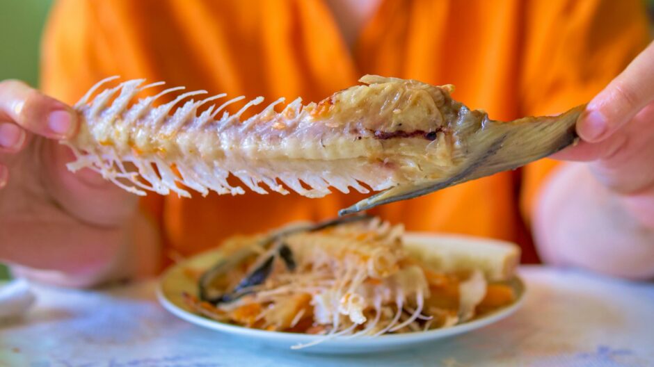 holding up a bare fish skeleton after finishing a seafood meal - displaying the spine and tail of a consumed whole fish above a messy plate