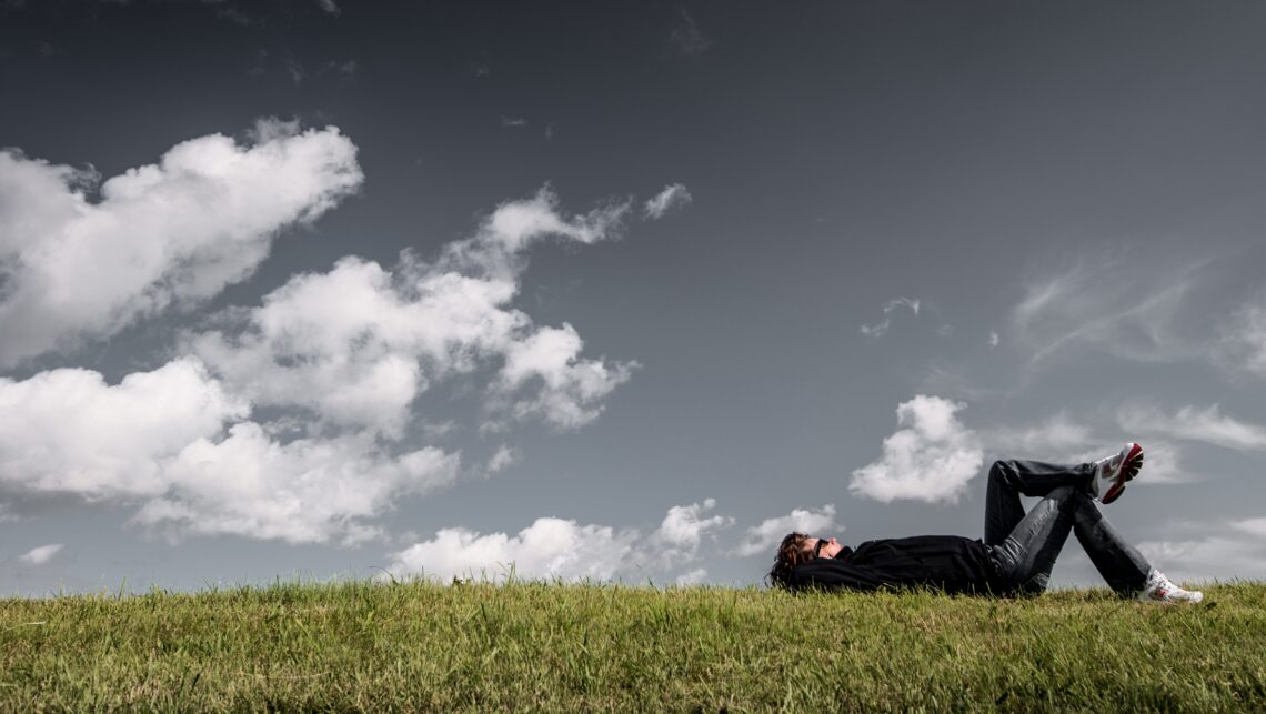 Man met sneakers ligt in het gras naar de wolken te kijken