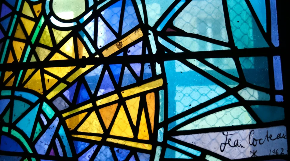 glas in lood in abstracte vorm, met geel en blauwe kleuren. Rechts onderin zijn een handtekening van Jean Cocteau en het jaartal 1962 in het glas geschilderd.